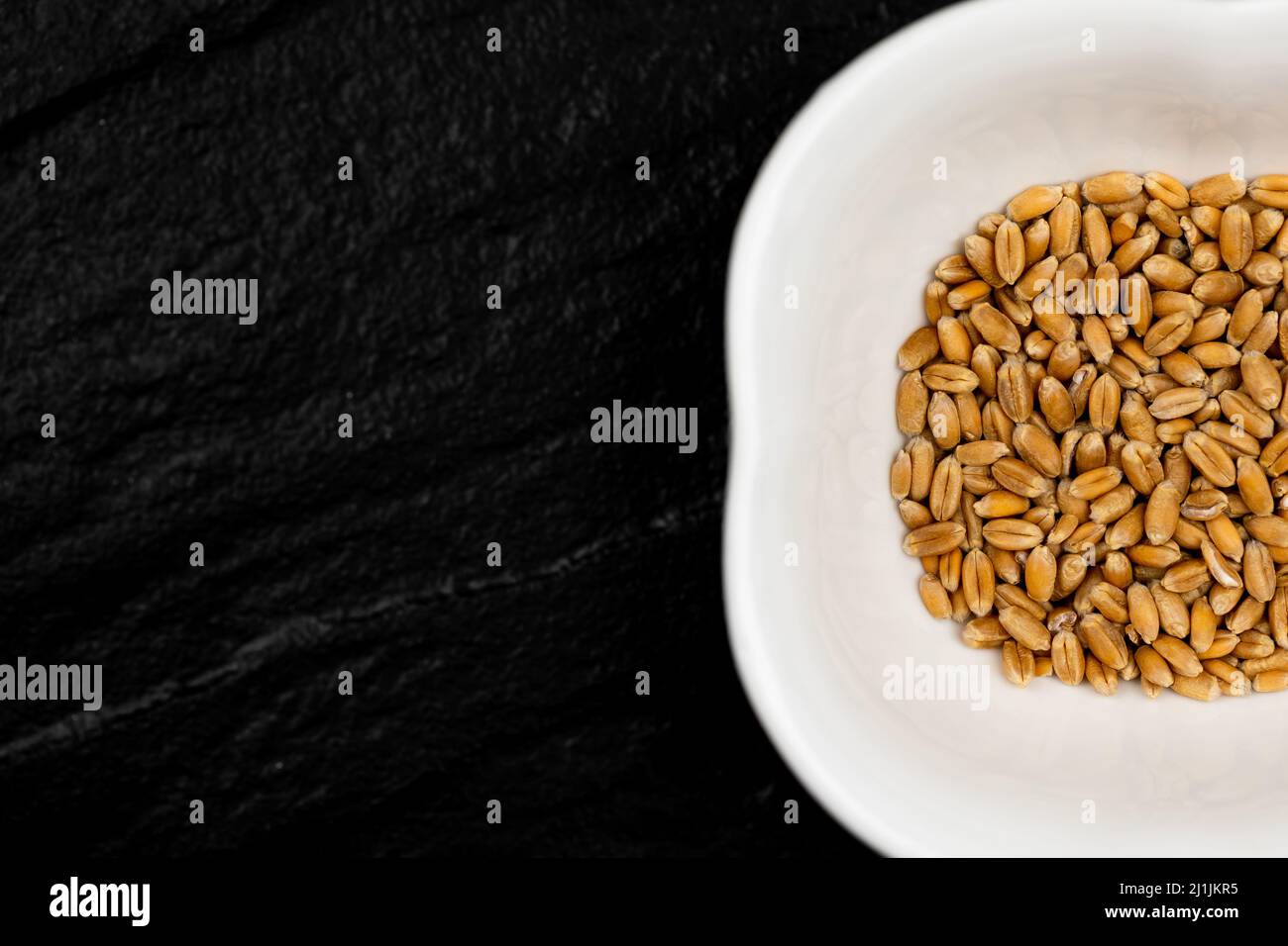 Vue de dessus du grain de blé dans un bol blanc sur une surface en pierre noire. Scène éclairée par une lumière artificielle douce. Banque D'Images