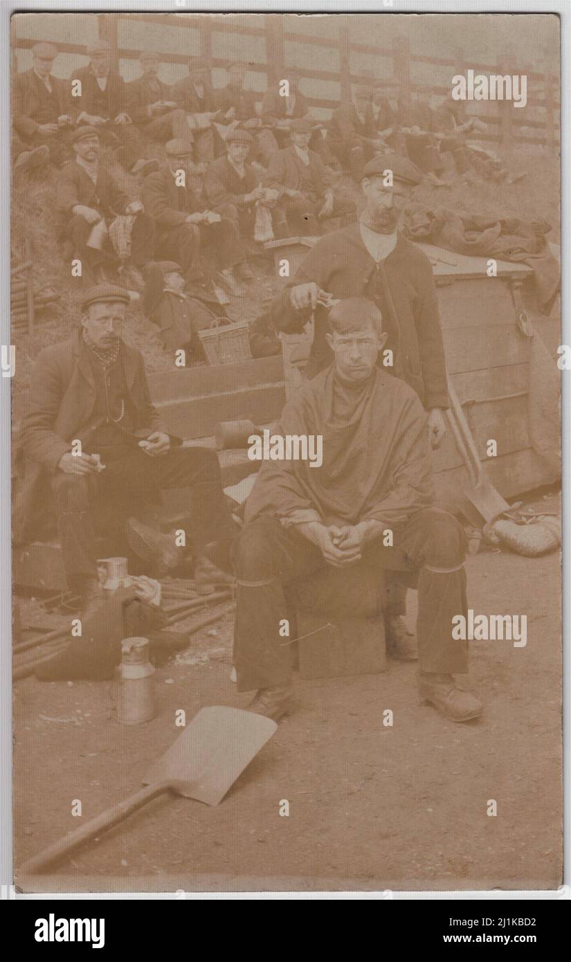 Coupe de cheveux, nourriture et boissons - naves au repos, début du 20th siècle. Groupe d'hommes assis sur une banque ayant de la nourriture et des boissons. Un homme est assis au premier plan en ayant ses cheveux coupés avec une paire de tondeuses. Des bêches, des ballons métalliques, des fladons, un panier et d'autres articles sont éparpillés Banque D'Images