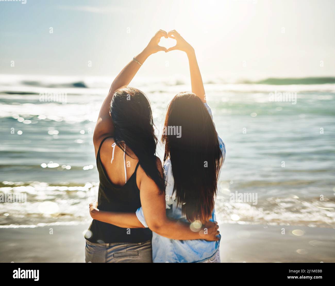 Nous partageons un amour pour les belles choses. Vue arrière de deux amis formant un coeur ensemble sur la plage. Banque D'Images
