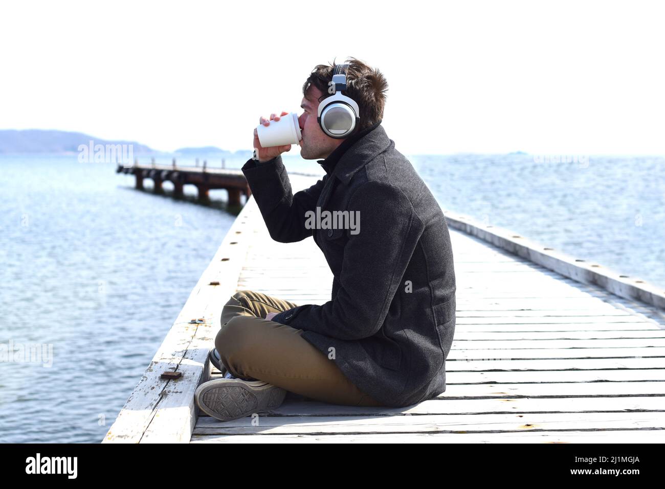 Jeune homme à l'écoute de musique et à boire du café, assis seul sur la jetée par une journée ensoleillée Banque D'Images