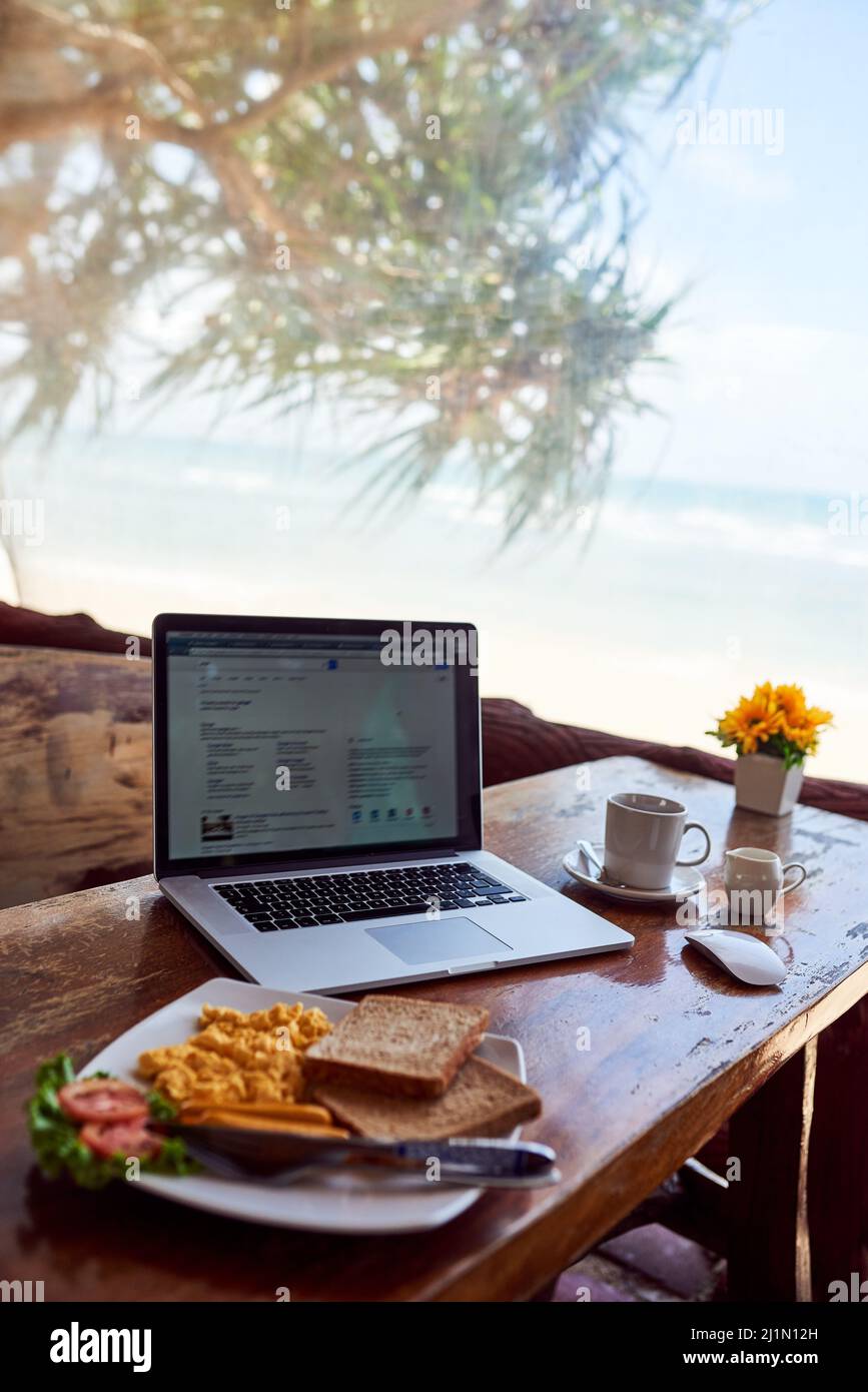 Réveillez-vous et brillez le long de la mer. Photo d'un ordinateur portable et petit déjeuner fraîchement préparé sur une table avec vue sur la plage en arrière-plan. Banque D'Images