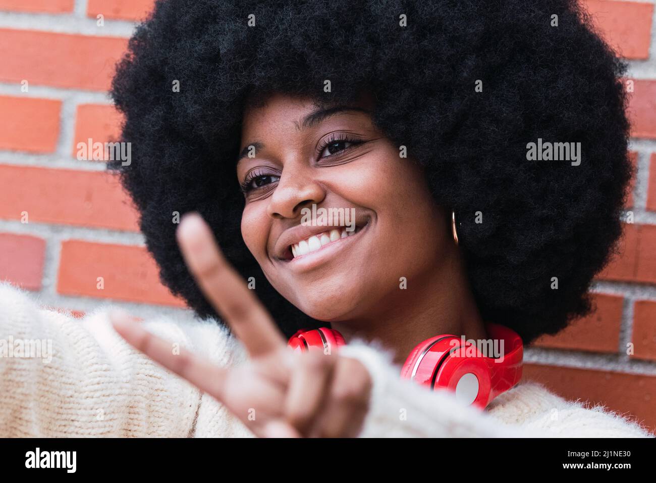 Gros plan portrait de la belle femme afro-américaine avec une coiffure afro-américaine, un pull blanc et des yeux fermés souriant tout en montrant le geste de paix Banque D'Images