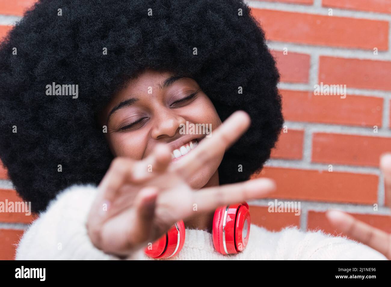 Gros plan portrait de la belle femme afro-américaine avec une coiffure afro-américaine, un pull blanc et des yeux fermés souriant tout en montrant le geste de paix Banque D'Images