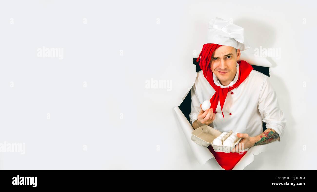Un homme beau avec des dreadlocks rouges le tient dans ses œufs de poulet de main. Jeune cuisinier confiant en chemise blanche et cravate rouge tenant les oeufs de poulet, peeking th Banque D'Images