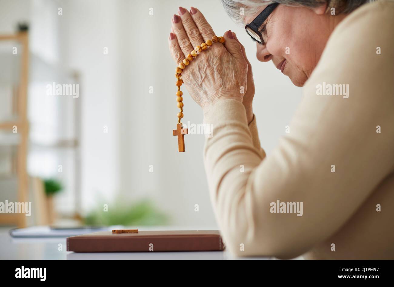 Femme chrétienne sénior assise à table avec la Sainte Bible, tenant rosaire et priant Banque D'Images