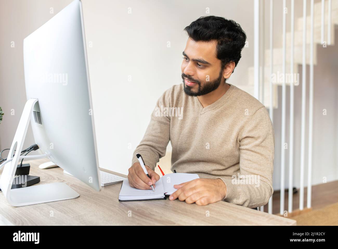 Homme indien positif concentré pensant, prenant des notes tout en étant assis à la table avec l'ordinateur à la maison. Auteur écrivant l'idée pour son livre, étudier en ligne. Concept d'apprentissage en ligne Banque D'Images