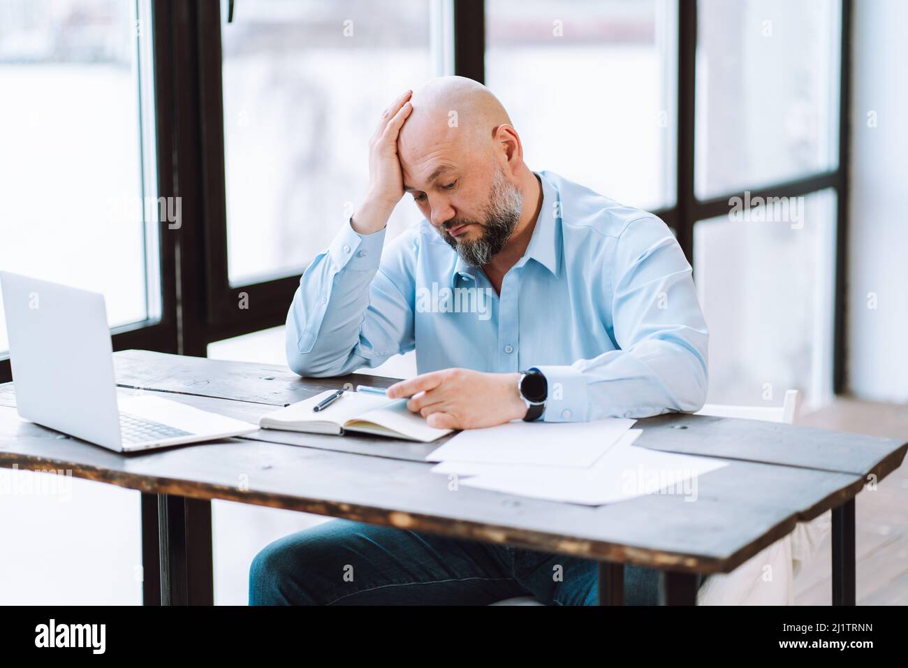 Portrait d'un bald d'âge moyen fatigué portant une chemise bleue, assis à un bureau en bois près de documents, ordinateur portable au bureau. Banque D'Images