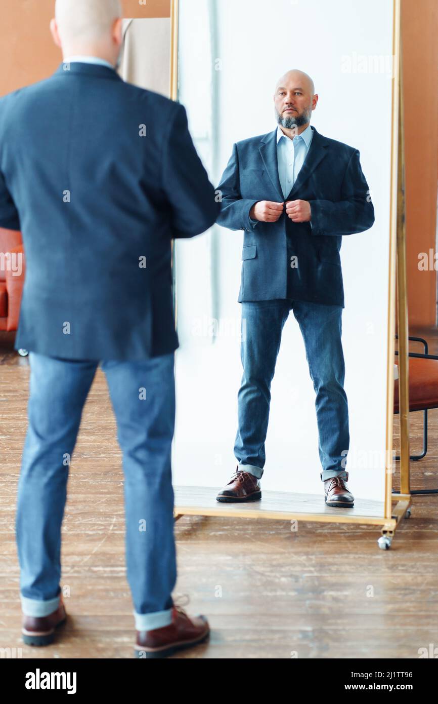 Portrait d'un homme bald d'âge moyen plein de confiance avec une barbe grise et une veste de costume bleue, en regardant le reflet miroir. Banque D'Images