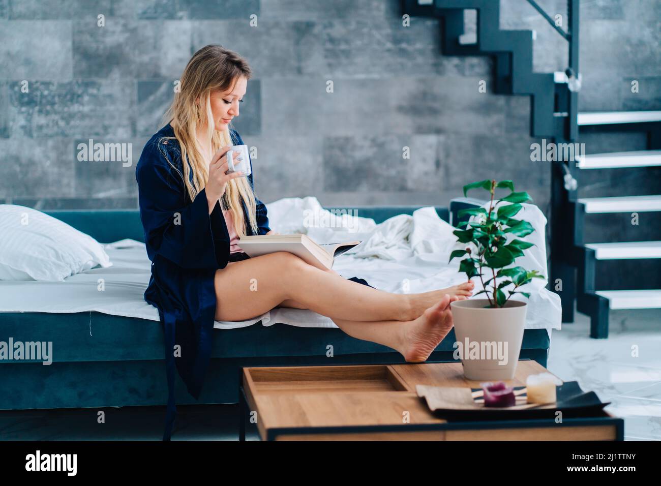 Vue latérale de la jeune femme merveilleuse assise avec des jambes croisées sur le canapé, portant robe de chambre, livre de lecture, tenir une tasse. Banque D'Images