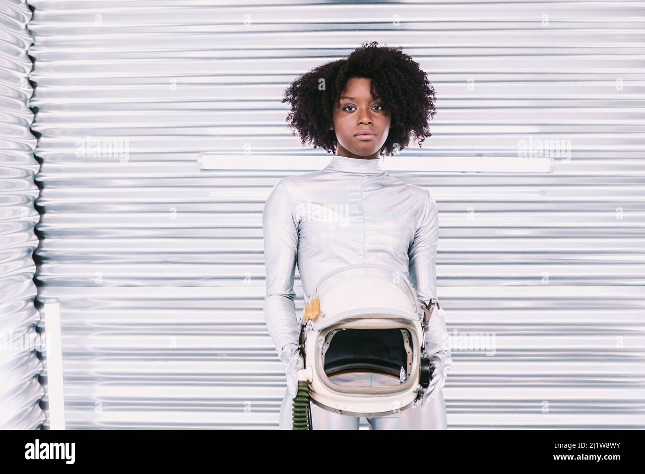Charmante femme afro-américaine avec casque en costume d'astronaute blanc regardant la caméra tout en se tenant dans un vaisseau spatial blanc sur fond clair Banque D'Images