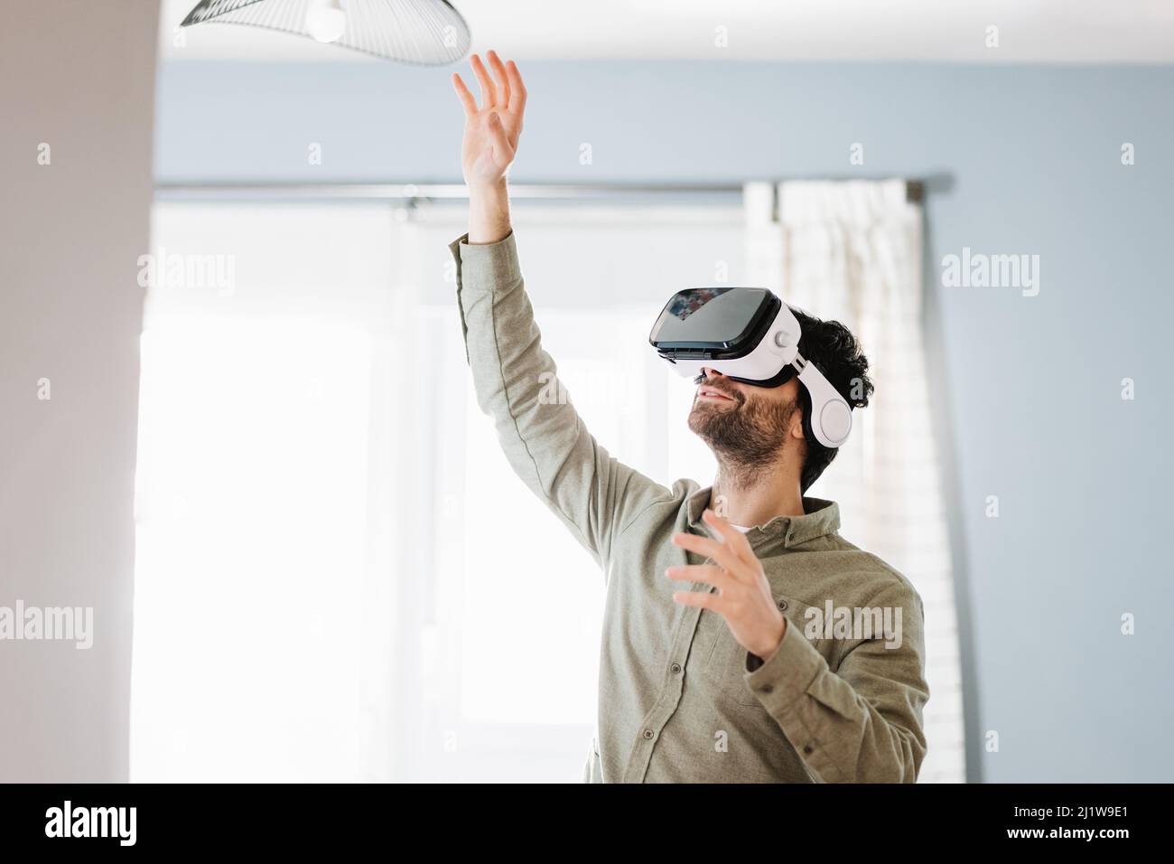 Jeune homme barbu avec des cheveux foncés dans des vêtements décontractés gestants et souriant tout en faisant l'expérience de la réalité virtuelle dans un casque moderne debout près de la fenêtre à Banque D'Images