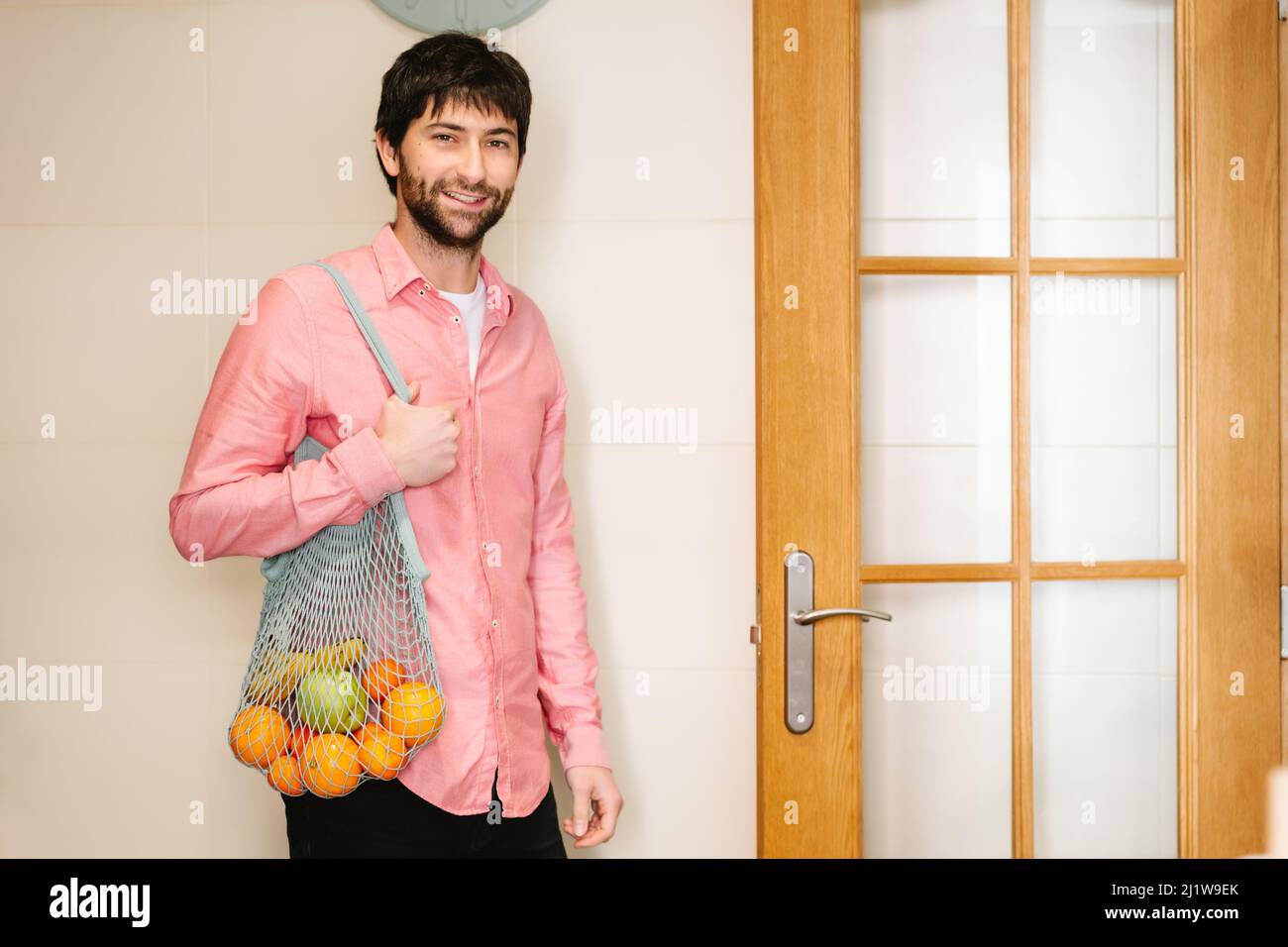 Positif jeune homme ethnique avec des cheveux foncés et une barbe dans des vêtements décontractés souriant et regardant l'appareil photo tout en se tenant dans la cuisine avec un sac en filet tissé avec Banque D'Images