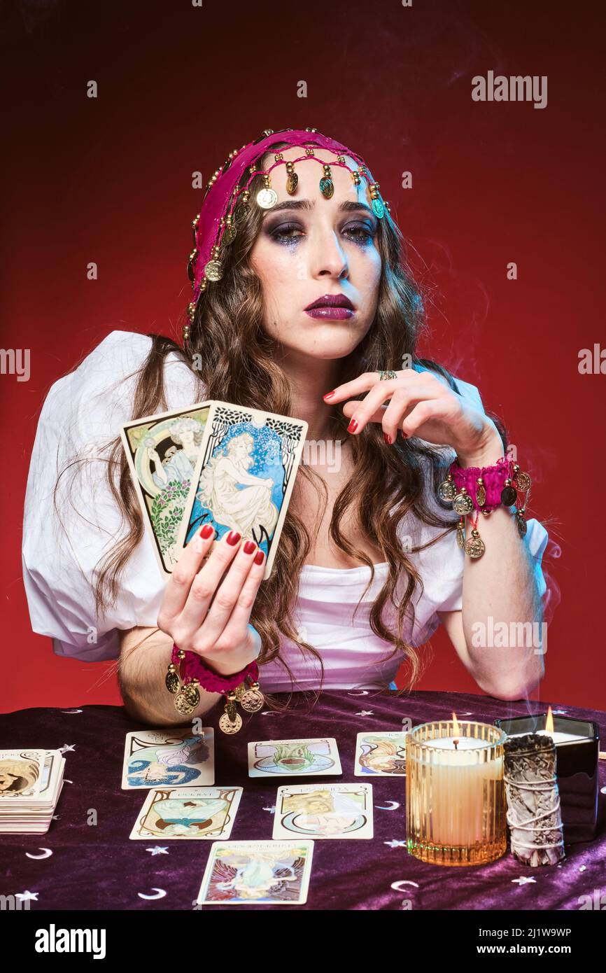Jolie femme apaisante avec maquillage assis à la table avec des cartes de tarot à la main contre les bougies allumées et regarder l'appareil photo Banque D'Images