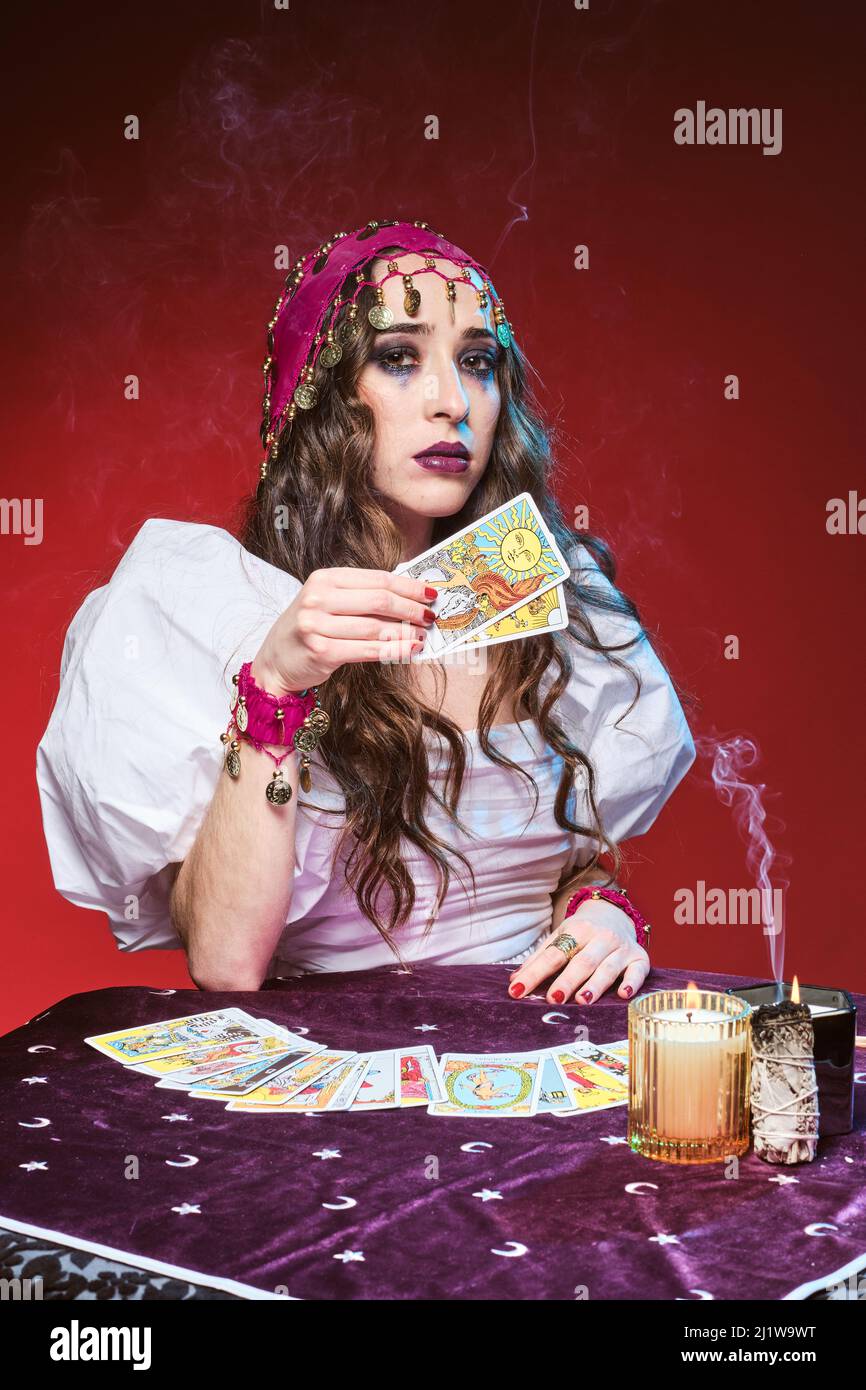 Jolie femme apaisante avec maquillage assis à la table avec des cartes de tarot à la main contre les bougies allumées et regarder l'appareil photo Banque D'Images