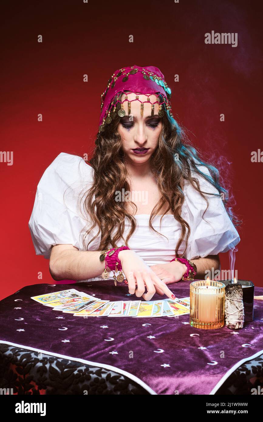 Jolie femme apaisante avec maquillage assis à la table avec des cartes de tarot contre les bougies allumées et regardant la table Banque D'Images