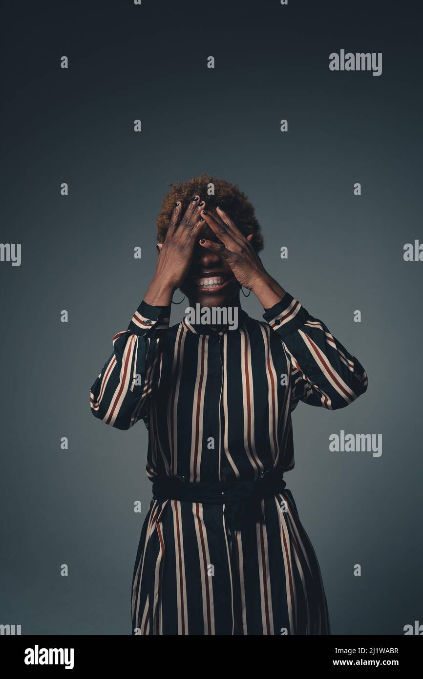 Une femme afro-américaine expressive riant largement sur une blague drôle et couvrant les yeux avec les mains dans un studio sombre Banque D'Images