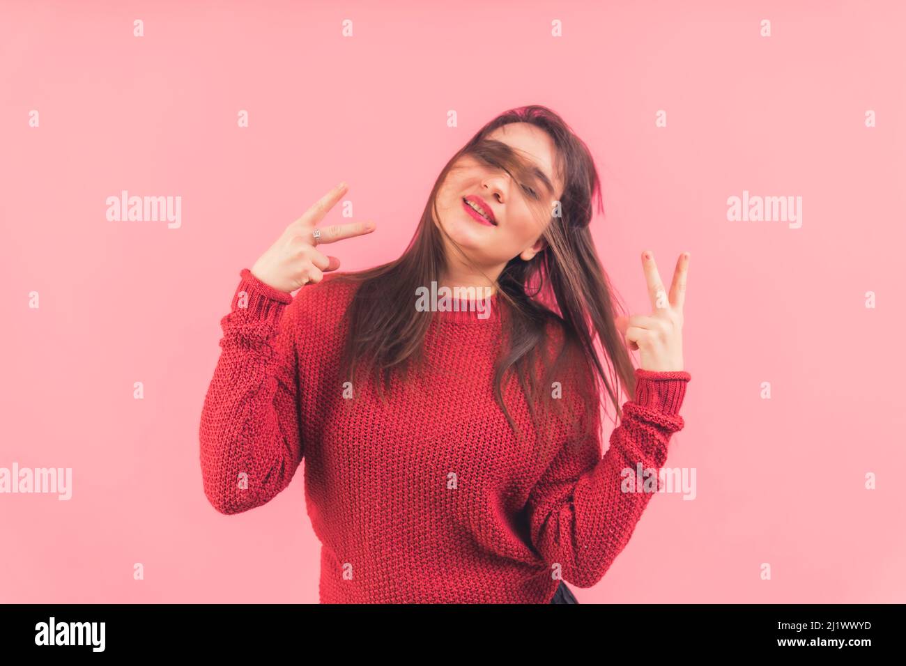 Jeune femme caucasienne avec des cheveux bruns soufflés au vent portant un chandail tricoté rouge tout en montrant le V-signe avec les deux mains sur fond rose. Photo de haute qualité Banque D'Images
