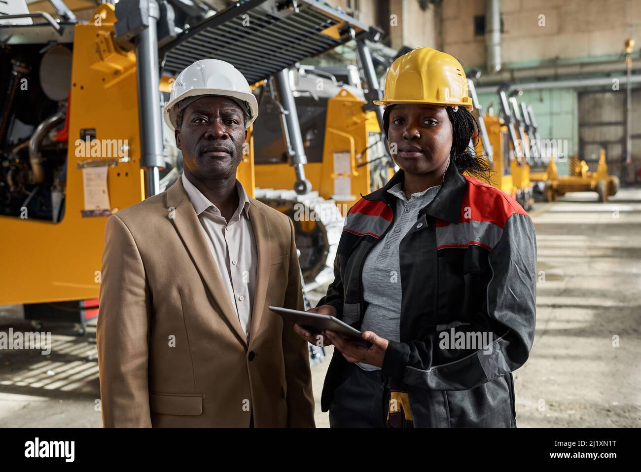 Portrait d'un ingénieur africain avec une femme mécanicien debout en usine et regardant l'appareil photo tout en travaillant en ligne sur une tablette pc Banque D'Images