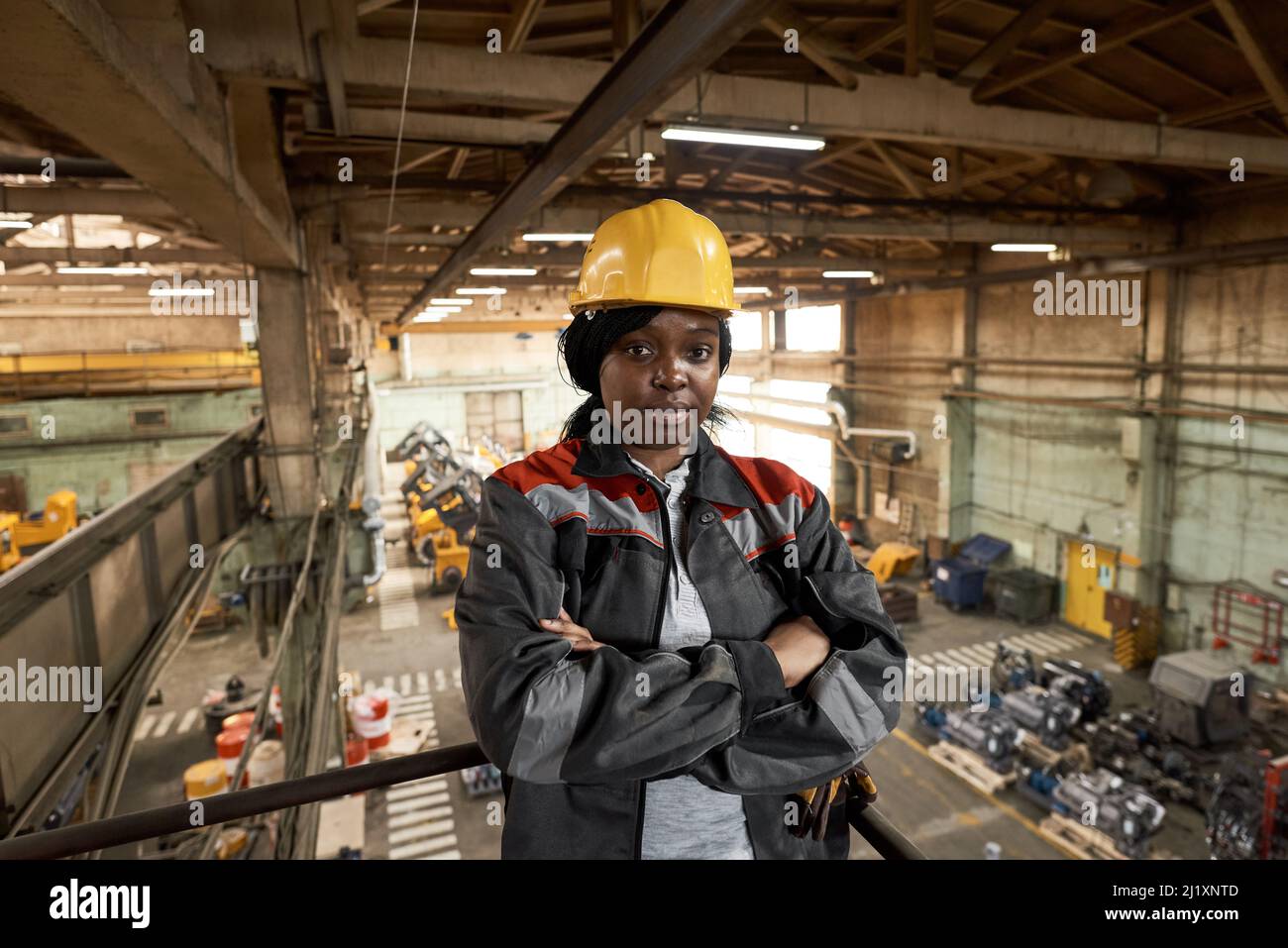 Portrait d'une travailleuse africaine en casque de travail et uniforme debout sur un balcon avec ses bras croisés et regardant la caméra Banque D'Images