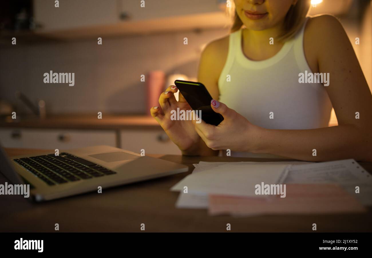 Une fille qui travaille dur travaille de la maison sur son ordinateur portable et vérifie son téléphone, et à côté d'elle est une pile de papiers Banque D'Images