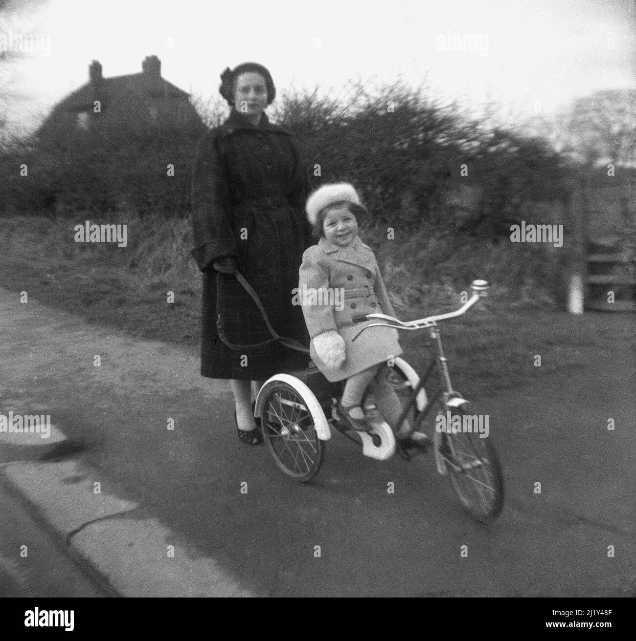 1950s, historique, à l'extérieur sur un trottoir de campagne, une mère avec sa fille assise sur son tricycle, Angleterre, Royaume-Uni. La dame tient une plomb attachée à l'arrière du tricycle, qui a été fait par TRIANG, un important fabricant britannique de jouets à base de métal pour enfants dans cette époque. Banque D'Images