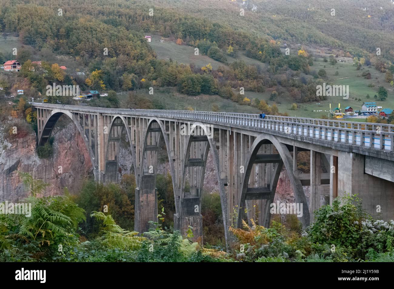 Pont de Tara au Monténégro avec couleurs d'automne et ciel bleu par une journée ensoleillée avec des collines et des maisons en arrière-plan Banque D'Images