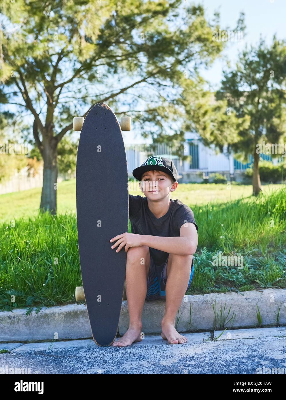 Apprendre à skate. Portrait complet d'un jeune garçon assis sur le trottoir avec sa longue planche. Banque D'Images