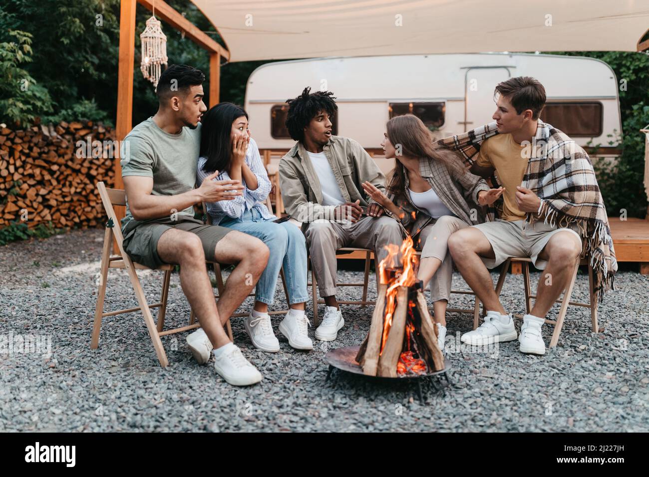 Divers jeunes amis assis près d'un feu de camp, racontant des histoires effrayantes à côté du camping-car pendant les vacances d'été Banque D'Images