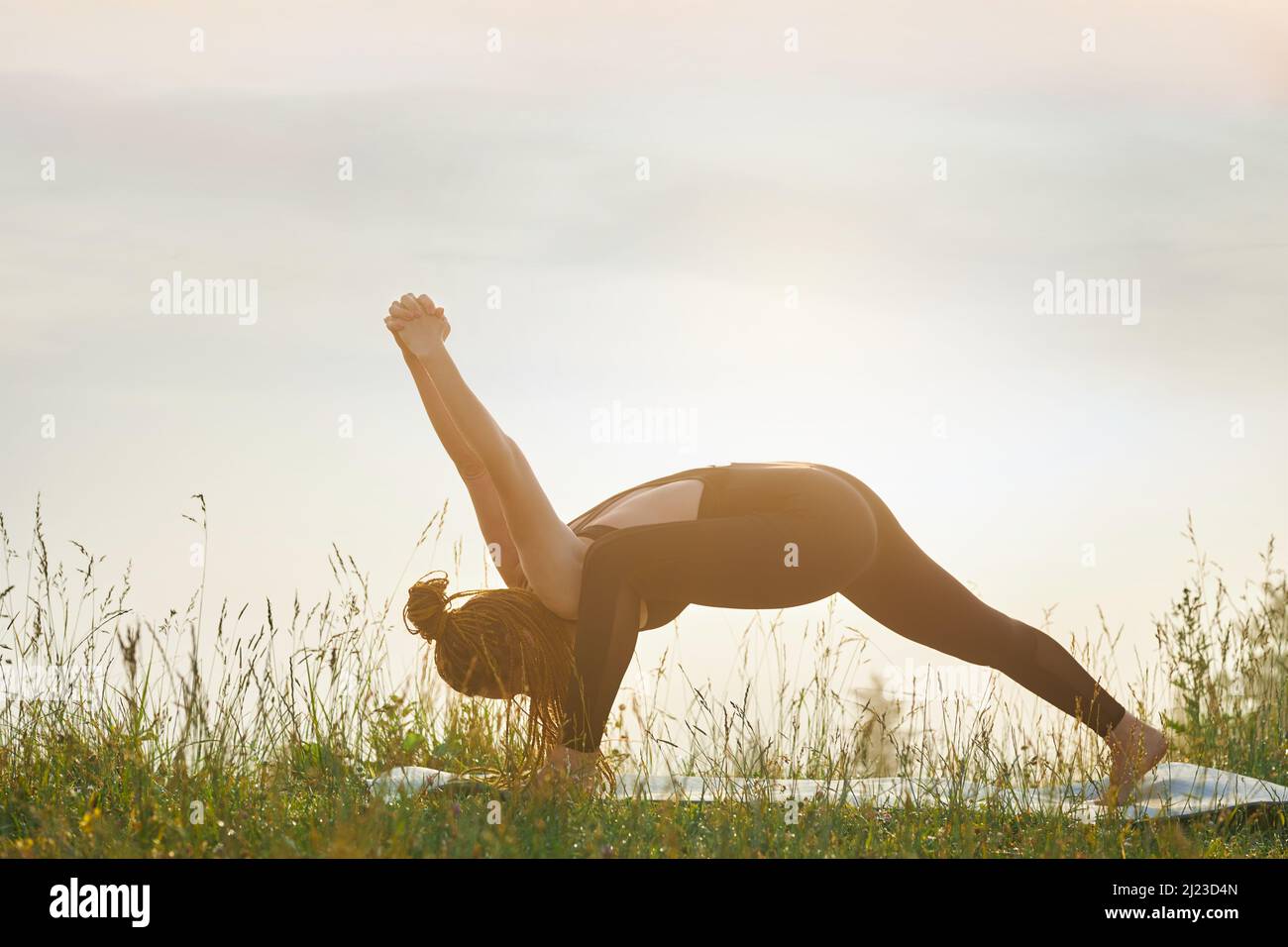 Vue latérale d'une femme flexible pratiquant le yoga. Femme sportive penchée vers l'avant du sol avec les mains levées en verrouillage sur le tapis de yoga dans la nature. Concept de nouvel âge. Banque D'Images