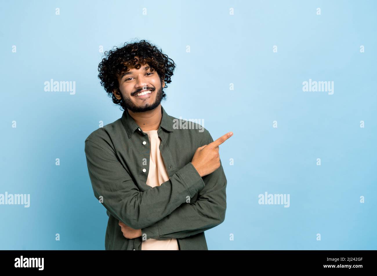Un homme à poil maurique indien ou arabe positif en tenue décontractée, pointe le doigt sur le côté de l'espace pour votre présentation, se tient sur un arrière-plan bleu isolé, regarde la caméra, souriant Banque D'Images