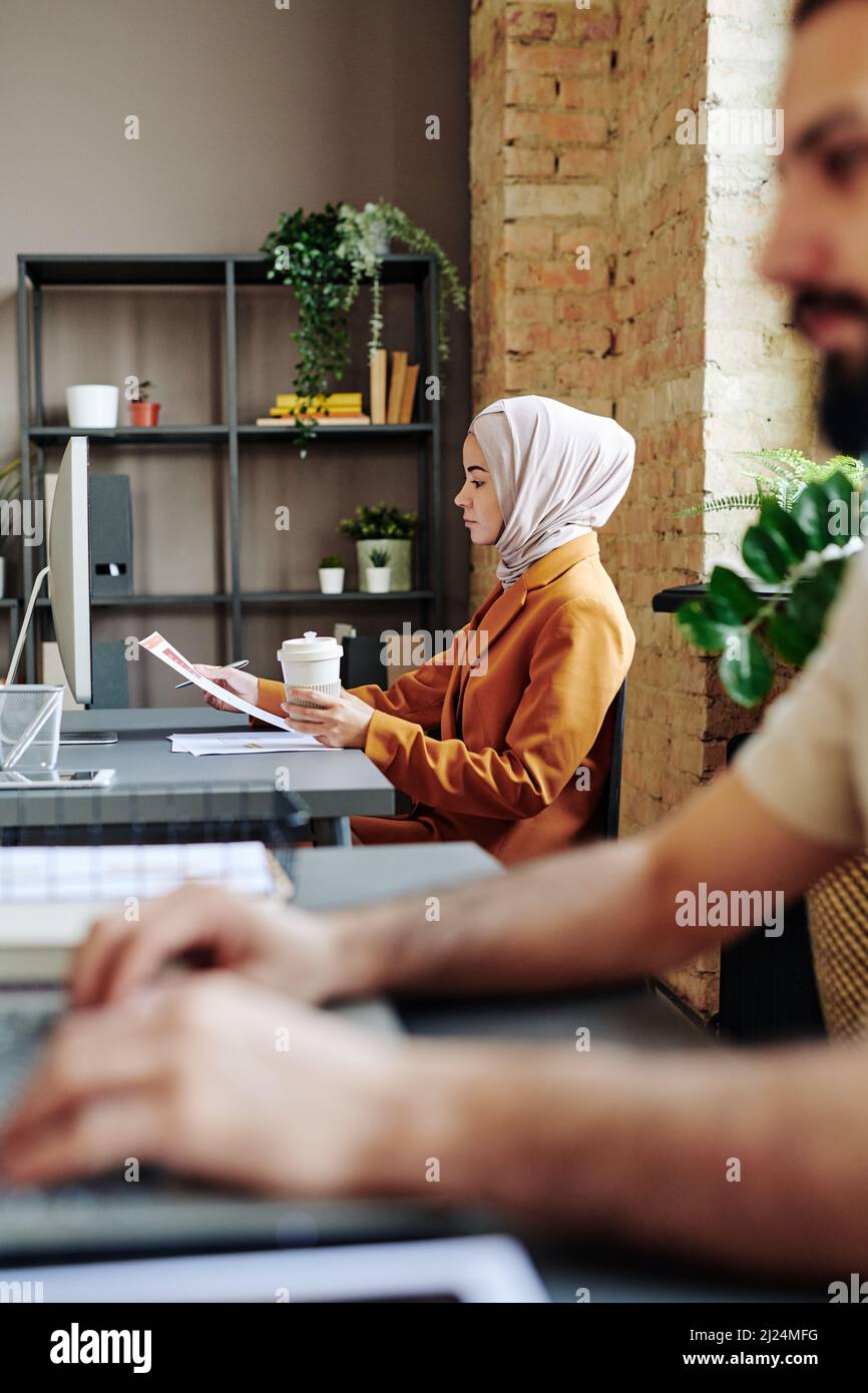 Jeune femme musulmane comptable en hijab et vêtements décontractés regardant à travers les journaux financiers tout en étant assise sur le lieu de travail Banque D'Images