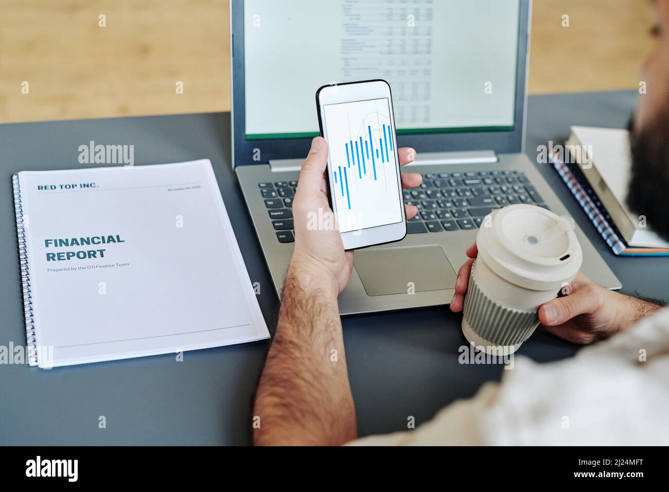 Jeune employé de bureau tenant un verre de café et un smartphone avec des tableaux financiers à l'écran tout en analysant les données par lieu de travail Banque D'Images