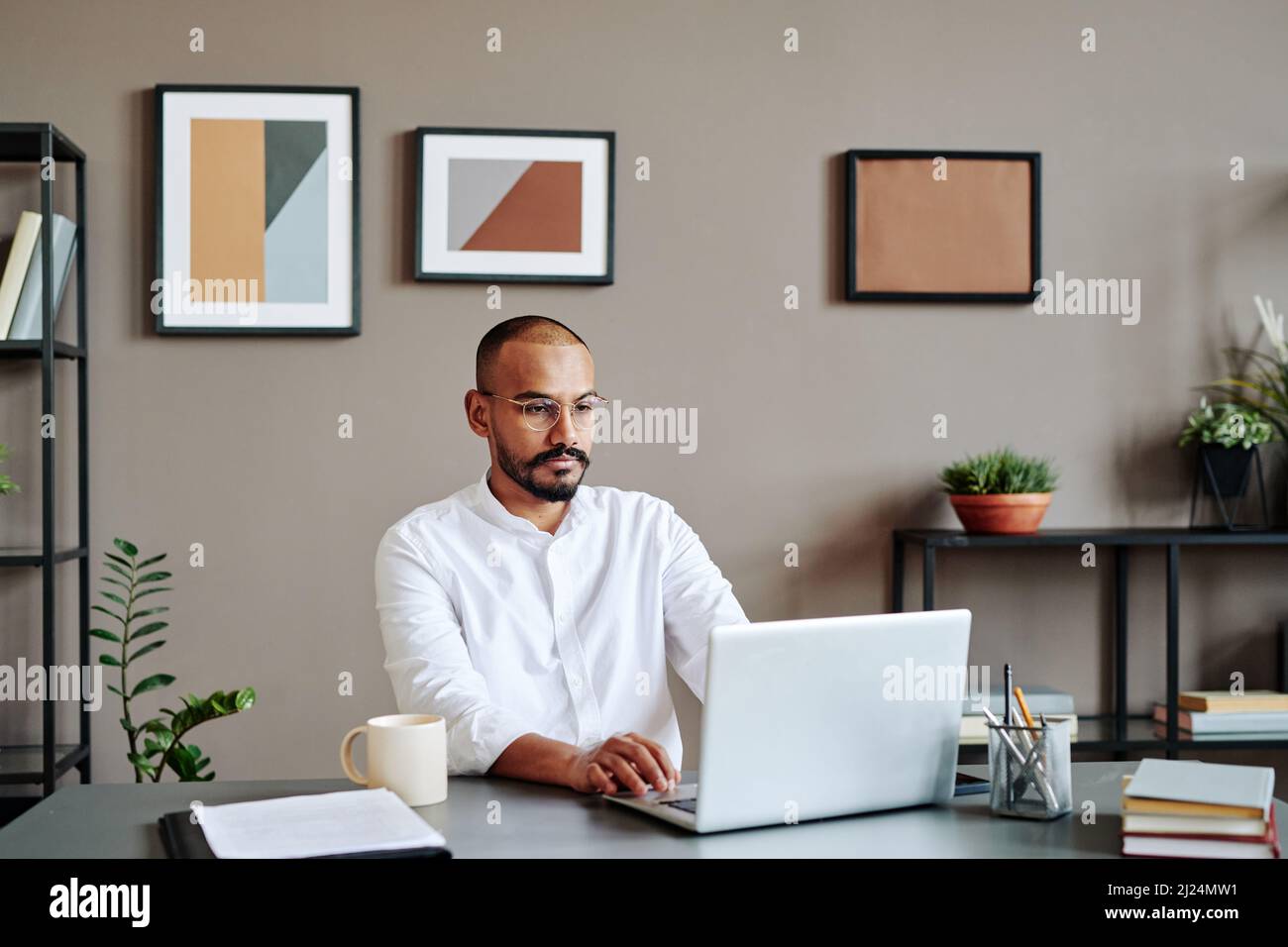 Jeune courtier moderne sérieux assis près du bureau et regardant l'écran d'ordinateur portable tout en analysant l'information financière en ligne Banque D'Images