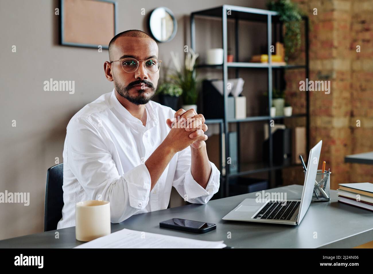 Jeune homme d'affaires contemporain qui garde ses mains jointes devant lui tout en étant assis sur son lieu de travail avec des gadgets Banque D'Images