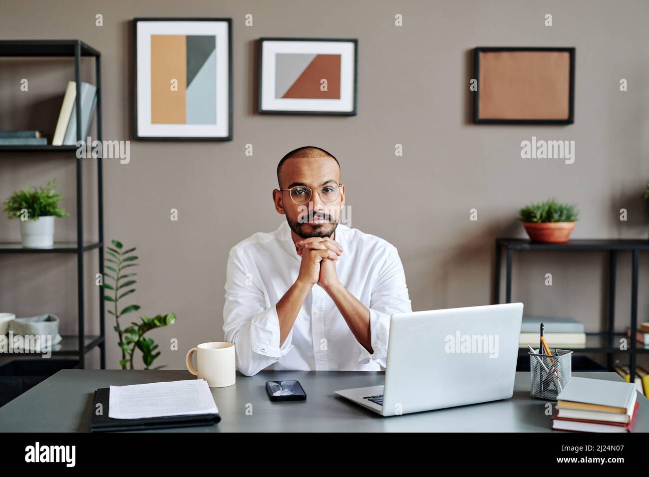 Jeune homme d'affaires musulman sérieux en chemise blanche, assis sur le lieu de travail devant un ordinateur portable et regardant un appareil photo pendant une réunion numérique Banque D'Images