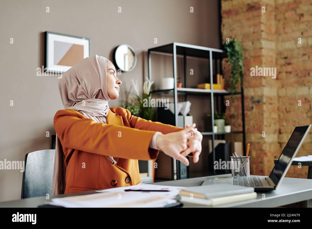 Jeune femme d'affaires musulmane contemporaine qui fait de l'exercice en s'asseyant sur le lieu de travail et regardant de côté au bureau Banque D'Images