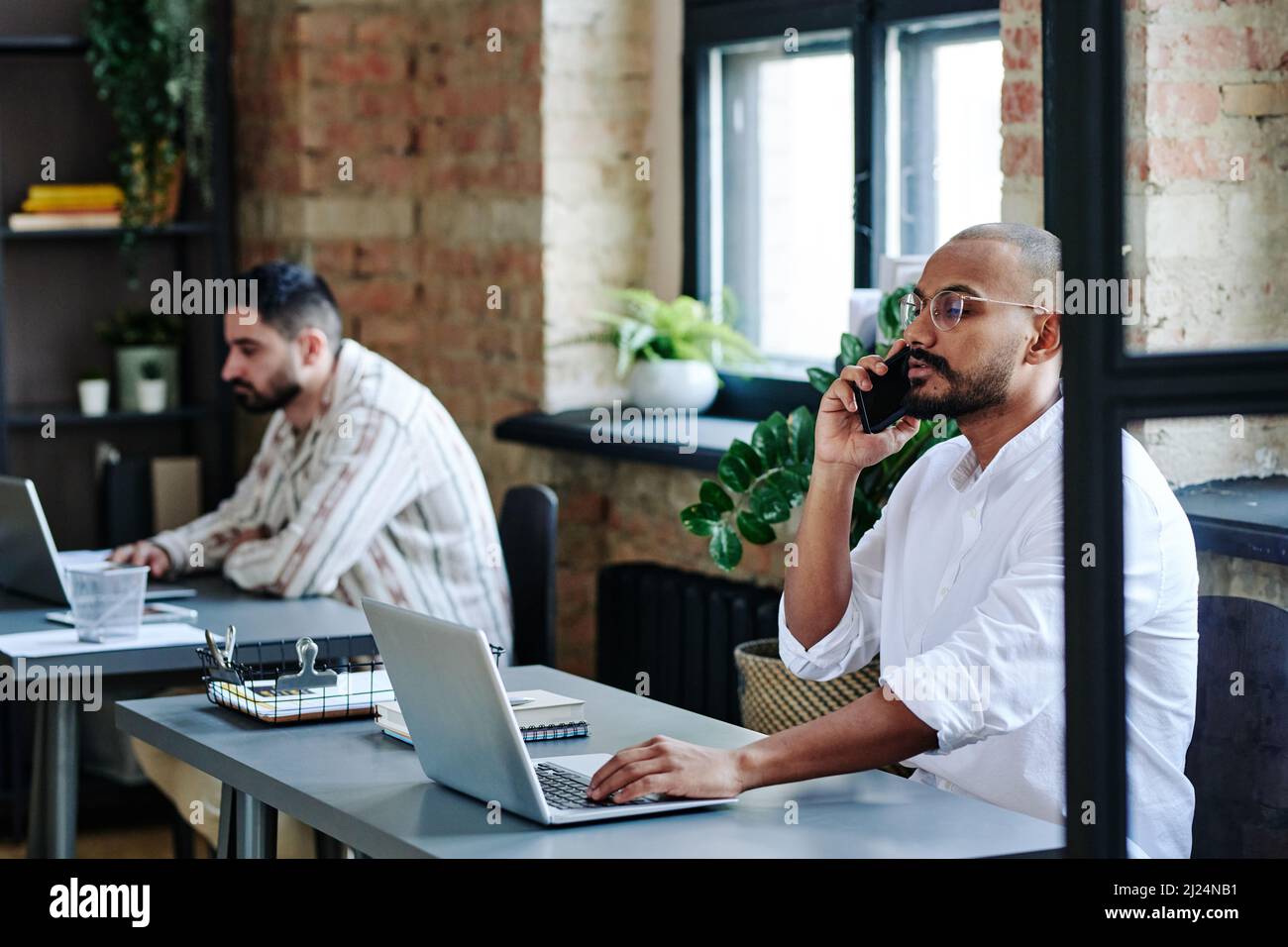 Jeune homme d'affaires confiant consultant le client sur un téléphone portable tout en étant assis près du bureau devant l'ordinateur portable contre un collègue de travail Banque D'Images