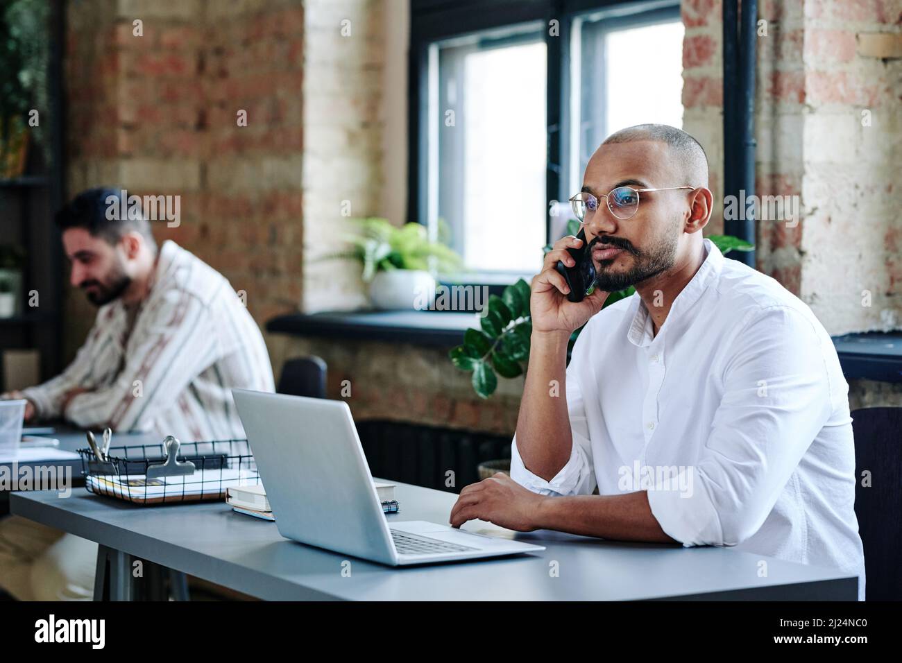 Jeune homme sérieux, employé de cols blancs, smartphone par oreille, regardant l'écran d'un ordinateur portable tout en lisant des informations en ligne au bureau Banque D'Images