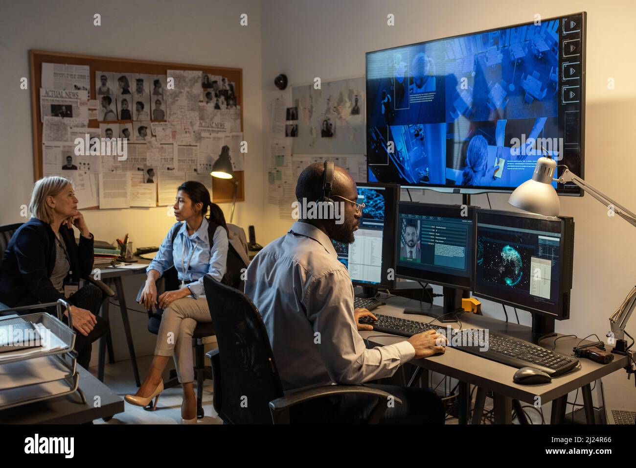Un jeune agent du FBI afro-américain assis devant des moniteurs d'ordinateur et devant un écran au bureau tout en regardant les enregistrements de caméras de sécurité Banque D'Images