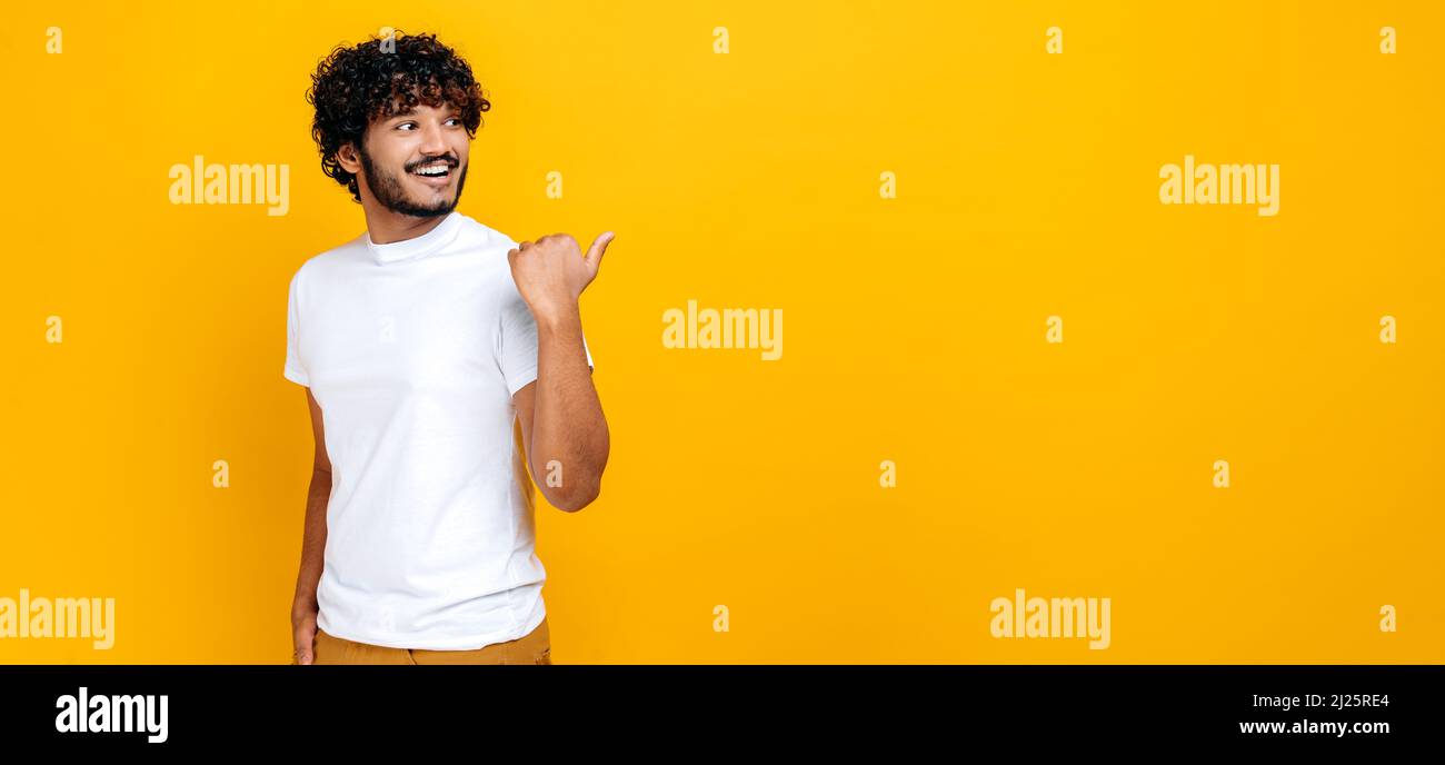 Photo panoramique d'un homme indien ou arabe aux cheveux bouclés et élégant, regardant et pointant du doigt sur le côté d'un espace de maquette vide, tout en se tenant sur un arrière-plan orange isolé, souriant Banque D'Images