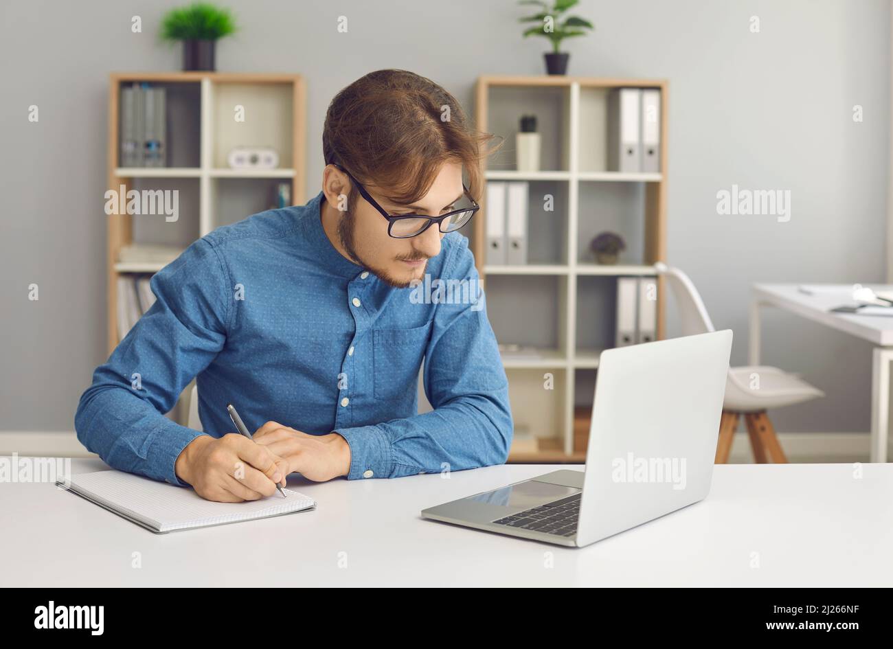 Un jeune homme concentré avec des lunettes prend des notes tout en travaillant avec un ordinateur portable à la maison. Banque D'Images