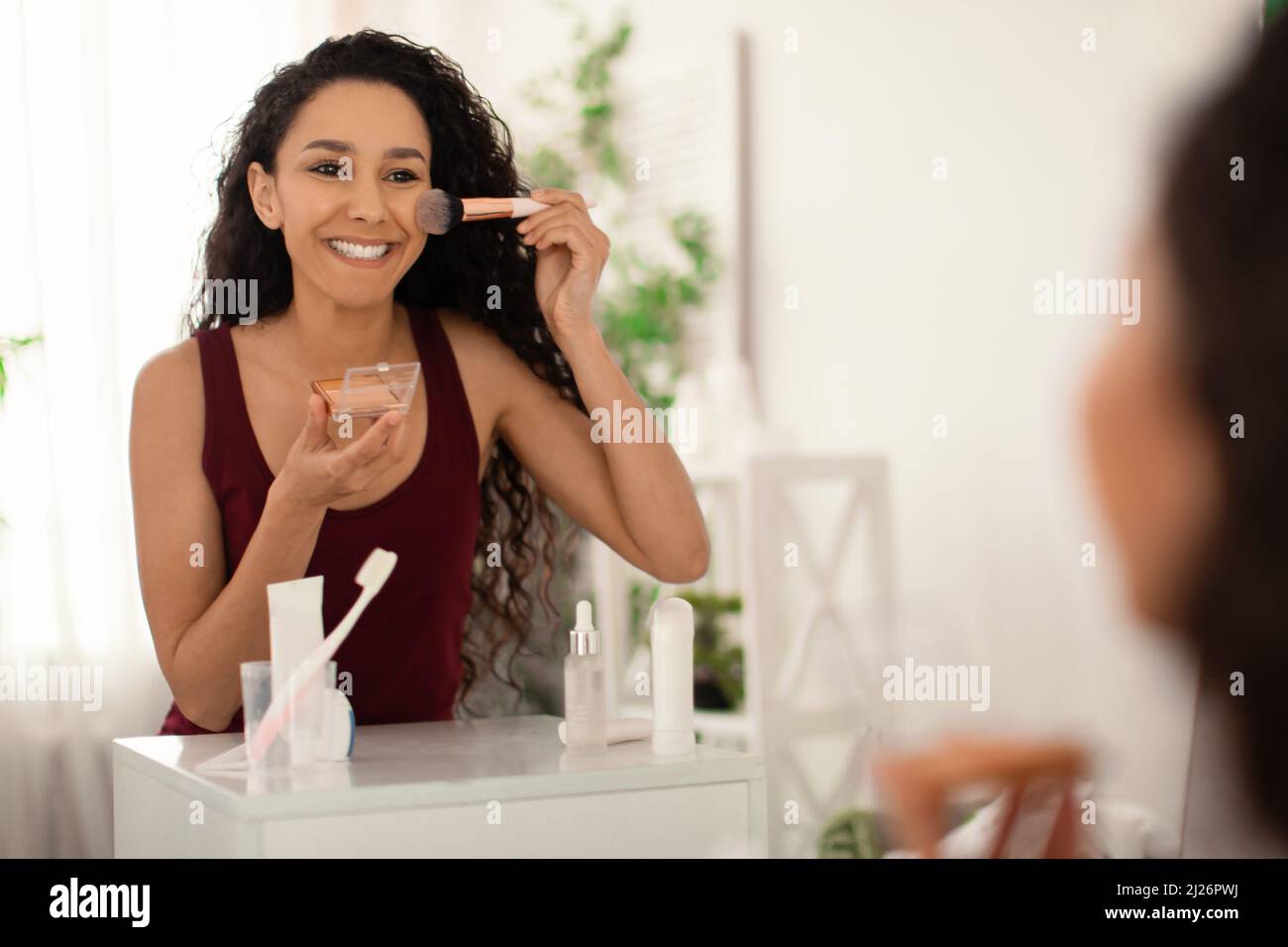Belle femme debout devant le miroir, tenant la brosse et appliquant le maquillage, le visage poudre ou bronzant dans la salle de bains Banque D'Images