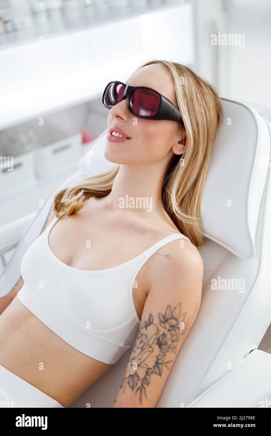 Femme blonde en lunettes de sécurité couchée sur le lit de jour et souriante en attendant la procédure de soin de la peau dans la clinique de cosmétologie Banque D'Images