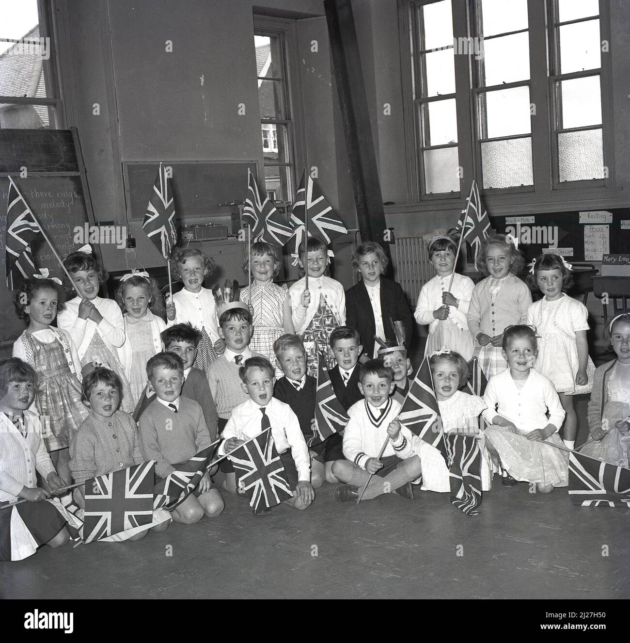 1965, historique, groupe d'enfants enthousiastes de l'école primaire assis dans une salle d'école tenant des drapeaux de Union Jack, Fife, Écosse, Royaume-Uni. Banque D'Images