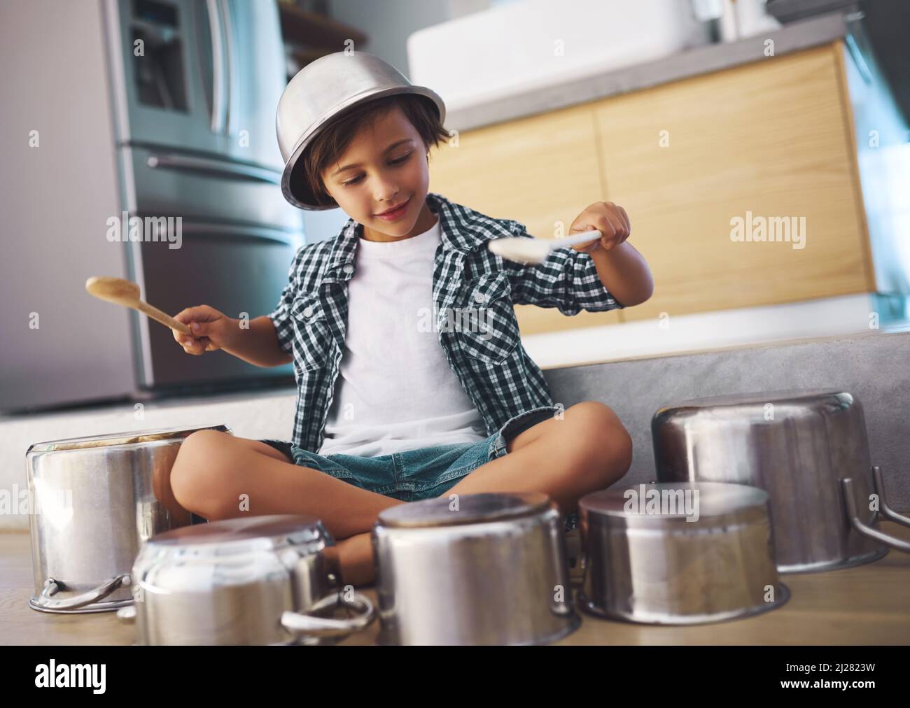 Déchaînement d'un tambour épique en solo. Photo d'un petit garçon heureux jouant des tambours avec des pots sur le sol de la cuisine tout en portant un bol sur sa tête. Banque D'Images