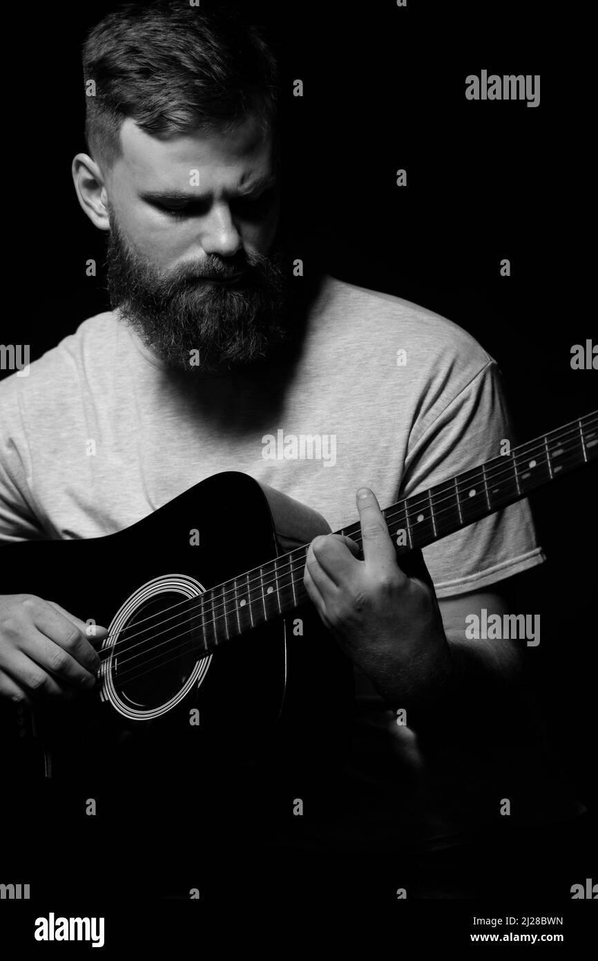 Le guitariste brutal barbu joue une guitare acoustique dans une pièce noire. Le concept d'enregistrement de musique, de répétition ou de spectacle en direct Banque D'Images