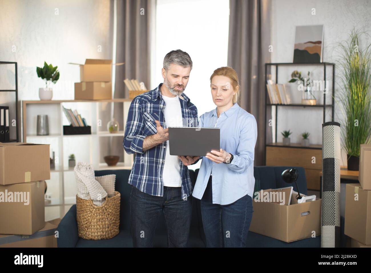 Couple d'âge mûr caucasien debout parmi les cardboxes et les différentes choses et en utilisant l'ordinateur portable, à la recherche de la conception et de faire des achats en ligne. La famille prend une pause lors de la dépose dans un nouvel appartement. Banque D'Images