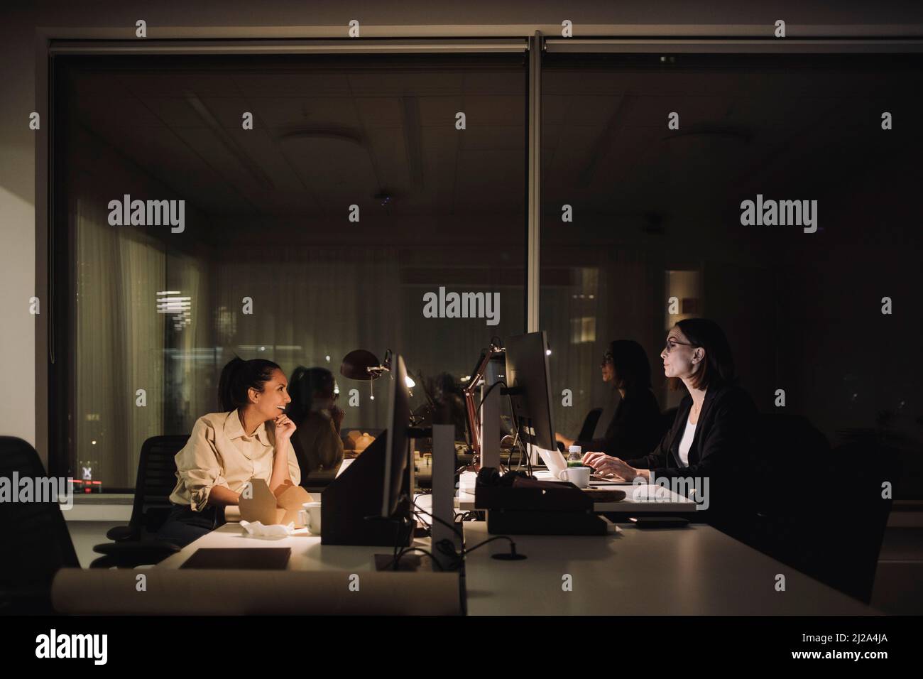 Femme d'affaires qui parle avec une collègue pendant qu'elle fait des heures supplémentaires sur le lieu de travail Banque D'Images