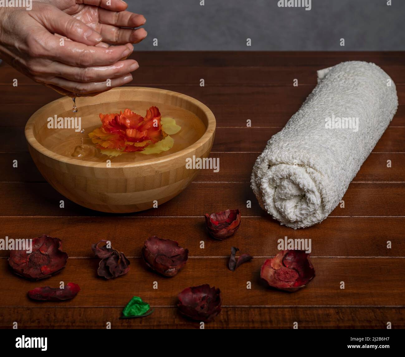 Les mains des femmes avec de l'eau s'écoulant dans un bol en bois avec une fleur jaune et rouge à l'intérieur, sur une table en bois avec une serviette enroulée Banque D'Images