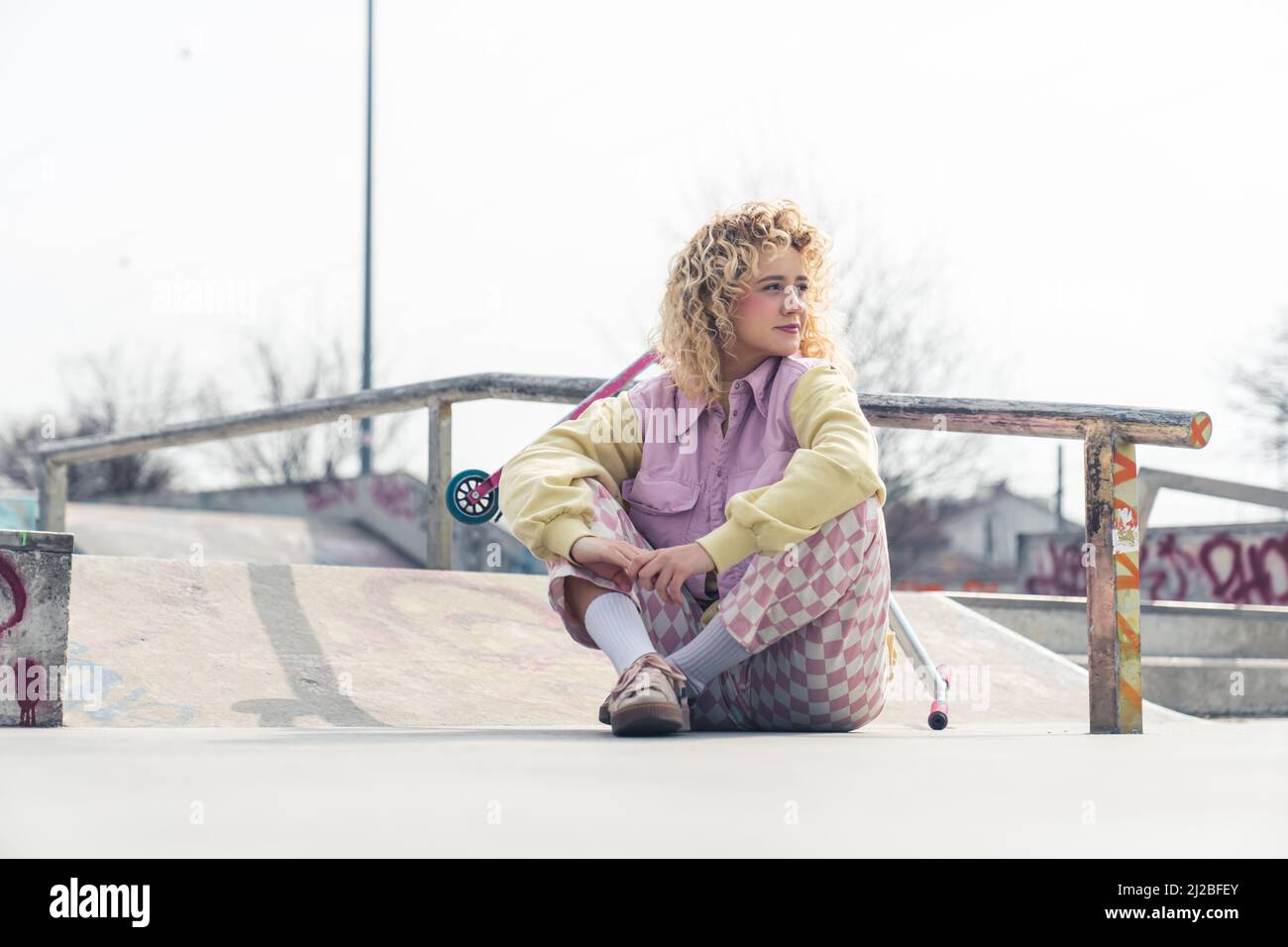 Jolie jeune femme blonde ruddy hippster est assise sur le sol de skateboard souriant, jambes croisées, regardant loin scooter rose sur l'arrière-plan plein espace de copie de plan. Photo de haute qualité Banque D'Images